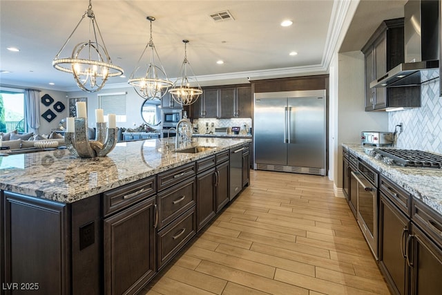 kitchen with sink, decorative light fixtures, built in appliances, a large island with sink, and wall chimney range hood