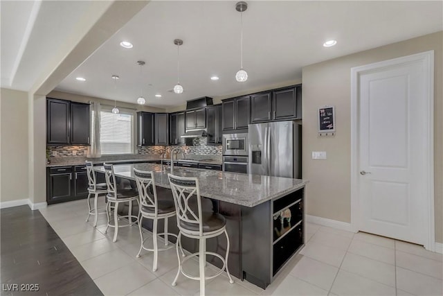 kitchen with appliances with stainless steel finishes, pendant lighting, an island with sink, decorative backsplash, and light stone counters