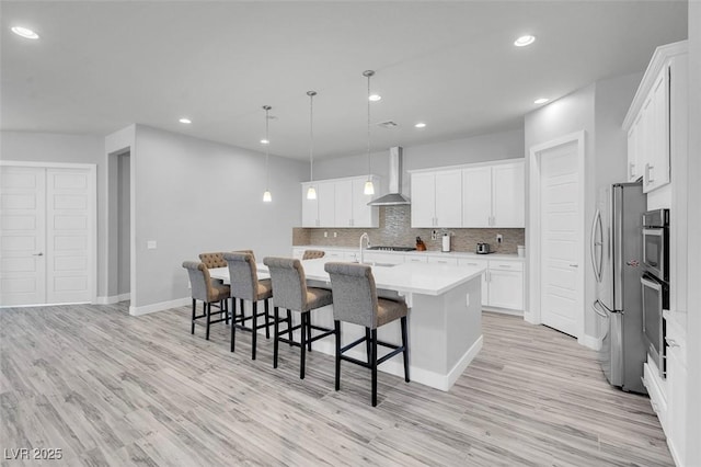 kitchen with pendant lighting, white cabinetry, an island with sink, stainless steel fridge, and wall chimney exhaust hood