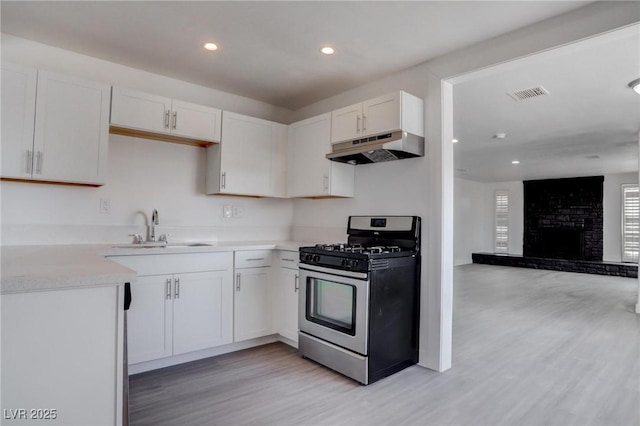 kitchen with sink, light hardwood / wood-style flooring, white cabinetry, stainless steel gas range oven, and a brick fireplace