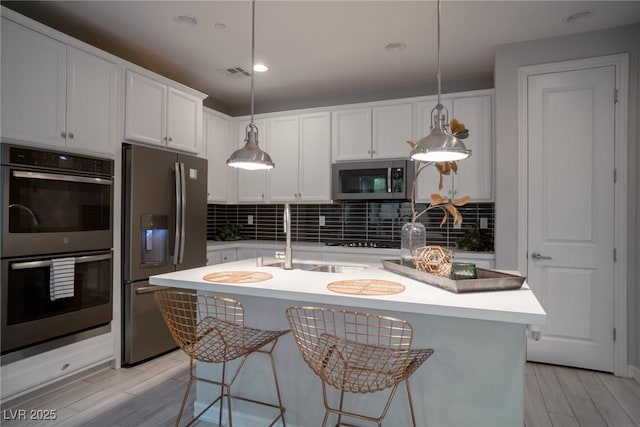 kitchen with a center island with sink, stainless steel appliances, hanging light fixtures, backsplash, and white cabinets