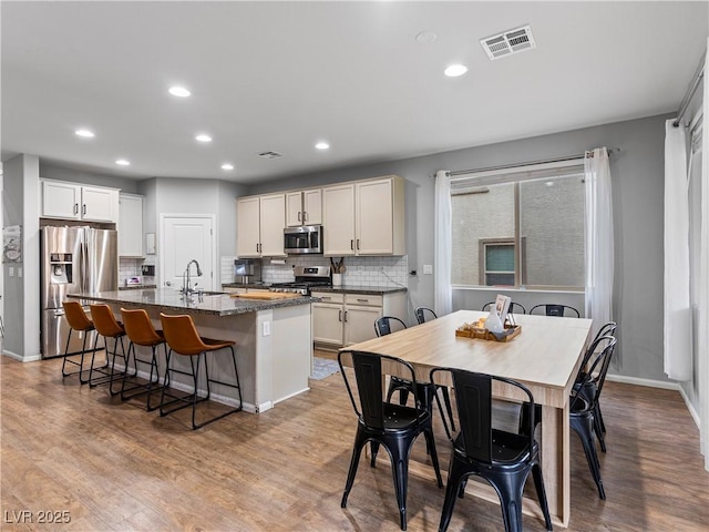 kitchen with tasteful backsplash, appliances with stainless steel finishes, a kitchen island with sink, dark stone counters, and light hardwood / wood-style floors