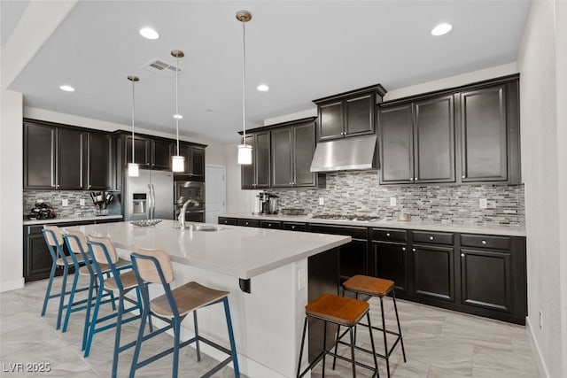 kitchen featuring light countertops, hanging light fixtures, a kitchen island with sink, stainless steel fridge, and under cabinet range hood