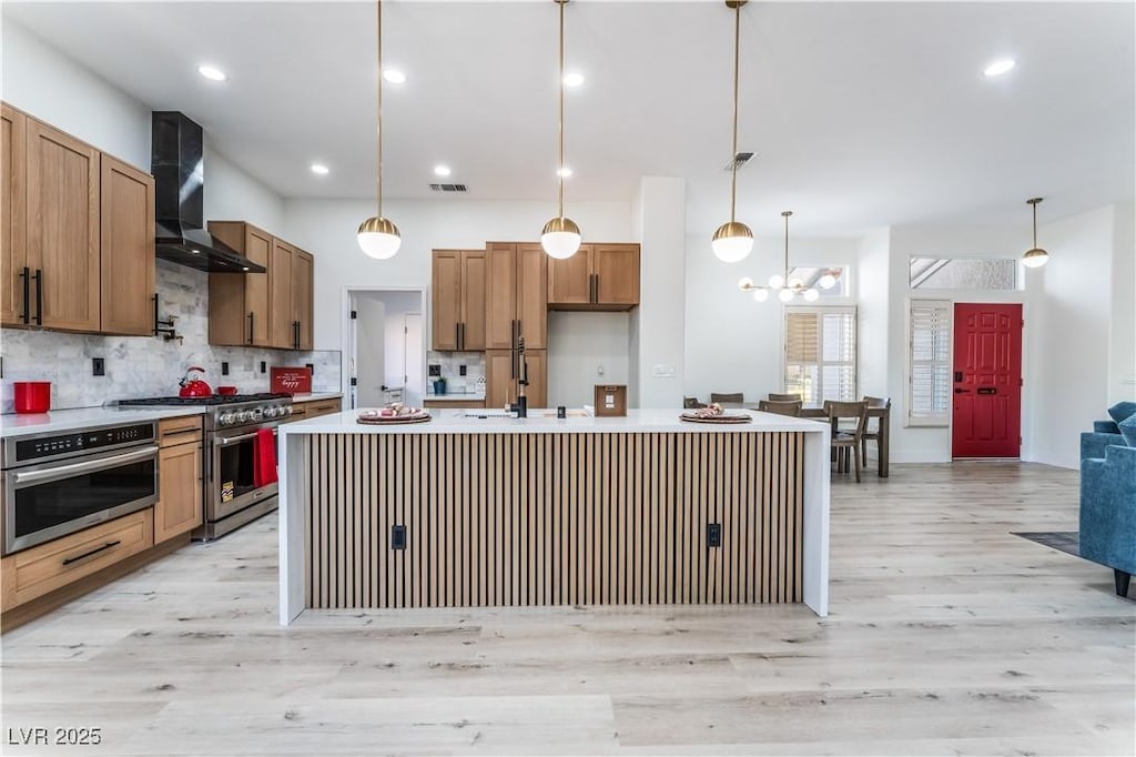 kitchen with sink, decorative light fixtures, an island with sink, stainless steel appliances, and wall chimney range hood