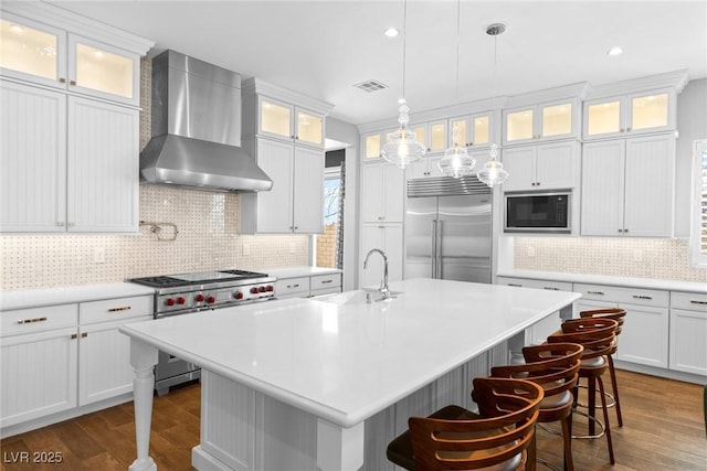 kitchen with an island with sink, sink, white cabinets, built in appliances, and wall chimney range hood