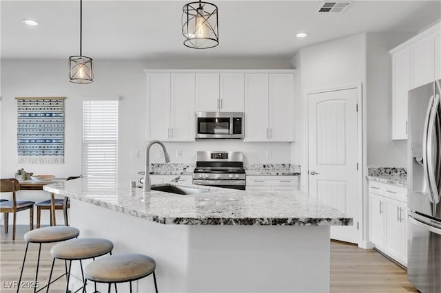 kitchen featuring appliances with stainless steel finishes, light stone counters, white cabinets, a center island with sink, and decorative light fixtures