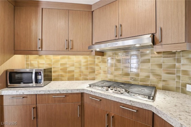 kitchen featuring light stone countertops, under cabinet range hood, stainless steel appliances, and decorative backsplash