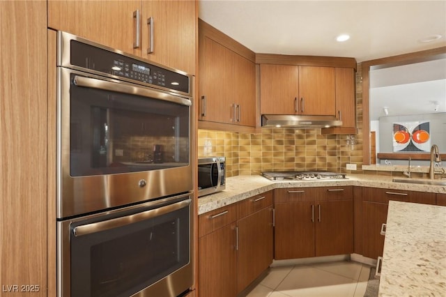 kitchen with light tile patterned floors, under cabinet range hood, a sink, appliances with stainless steel finishes, and decorative backsplash