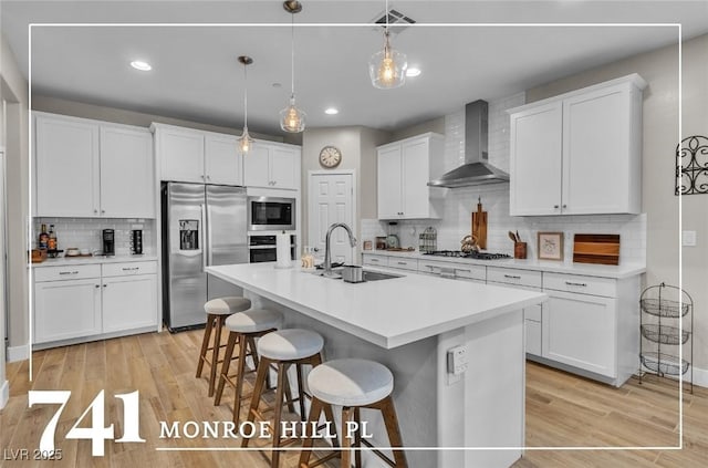 kitchen with wall chimney exhaust hood, sink, stainless steel appliances, a kitchen island with sink, and white cabinets