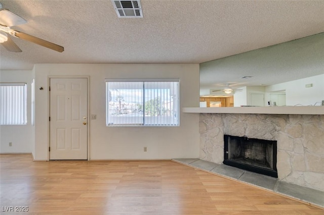 unfurnished living room with ceiling fan, a stone fireplace, a textured ceiling, and light wood-type flooring