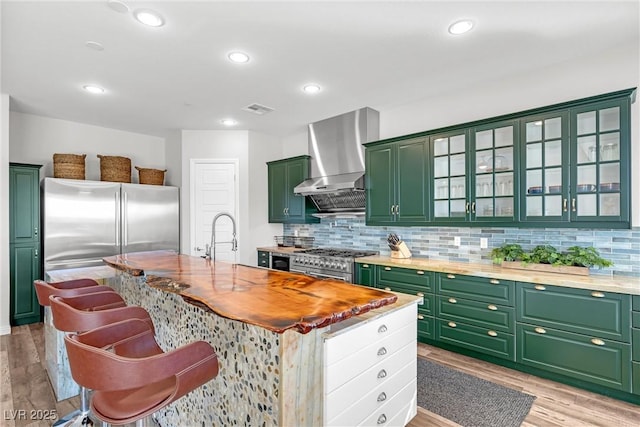 kitchen featuring high quality appliances, sink, butcher block counters, a center island with sink, and wall chimney exhaust hood