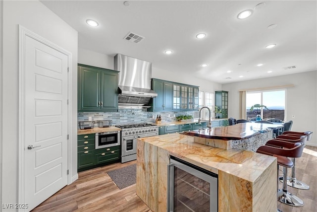 kitchen with a breakfast bar, beverage cooler, green cabinets, stainless steel appliances, and wall chimney exhaust hood