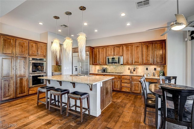 kitchen featuring appliances with stainless steel finishes, dark hardwood / wood-style floors, pendant lighting, light stone countertops, and a center island with sink