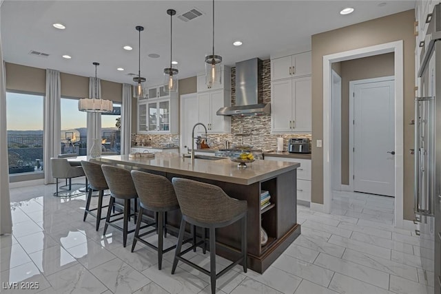 kitchen featuring marble finish floor, tasteful backsplash, a kitchen island with sink, a sink, and wall chimney exhaust hood