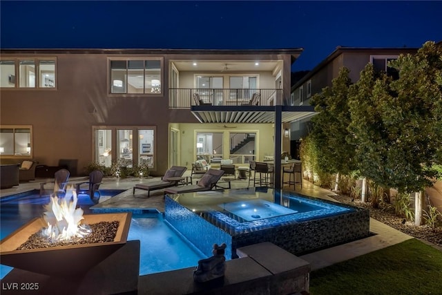 back of house at twilight featuring a balcony, stucco siding, an outdoor fire pit, and an in ground hot tub