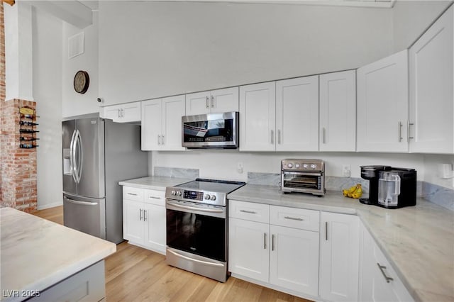 kitchen with light stone counters, stainless steel appliances, a towering ceiling, white cabinetry, and light wood-type flooring