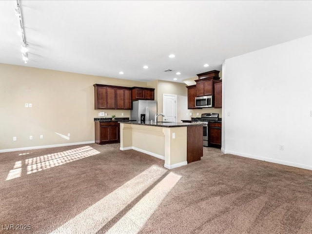 kitchen featuring light colored carpet, stainless steel appliances, baseboards, dark countertops, and a center island with sink