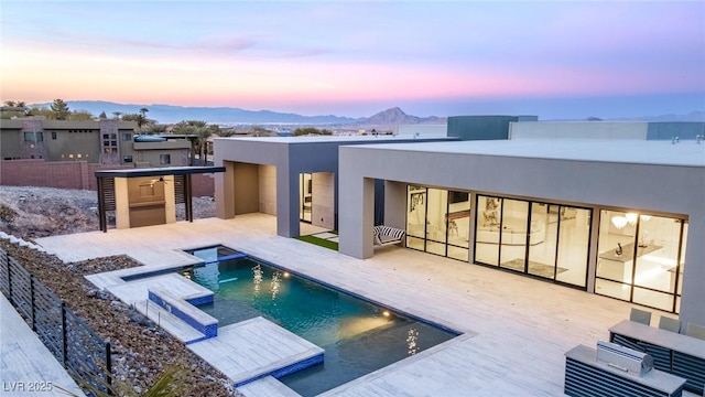 back of property at dusk with fence, a patio area, a pool with connected hot tub, a mountain view, and stucco siding