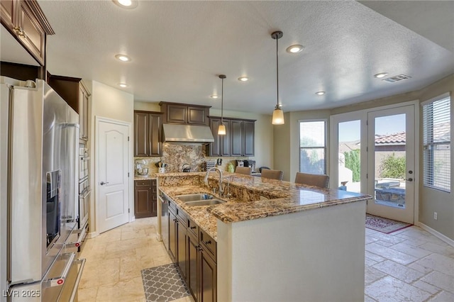 kitchen with sink, hanging light fixtures, stainless steel appliances, an island with sink, and decorative backsplash