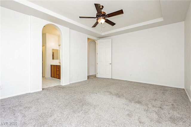 empty room with ceiling fan, arched walkways, light colored carpet, a raised ceiling, and baseboards