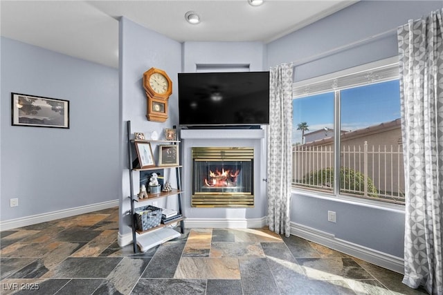 living room featuring stone tile flooring, a glass covered fireplace, and baseboards