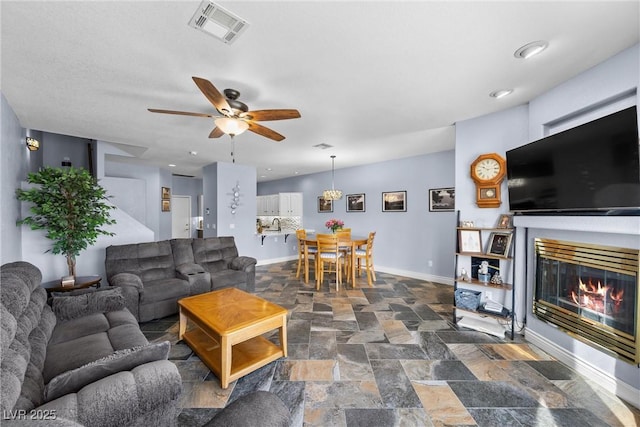 living area featuring a ceiling fan, baseboards, visible vents, stone finish flooring, and a glass covered fireplace