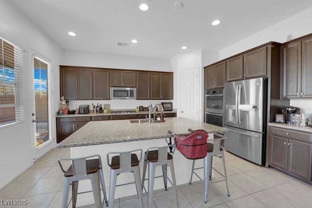 kitchen featuring a breakfast bar area, dark brown cabinetry, light stone counters, stainless steel appliances, and a center island with sink