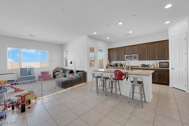 kitchen with light tile patterned flooring, a breakfast bar, light stone counters, dark brown cabinets, and a kitchen island with sink