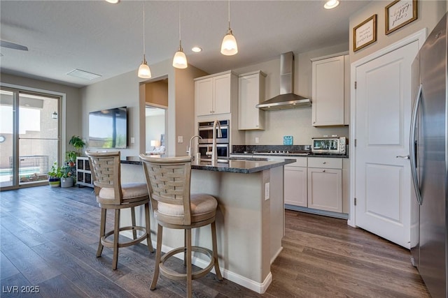 kitchen featuring a center island with sink, decorative light fixtures, white cabinets, wall chimney range hood, and a breakfast bar area