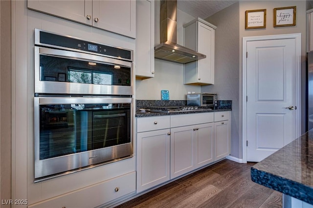 kitchen with dark stone countertops, appliances with stainless steel finishes, wall chimney range hood, dark hardwood / wood-style flooring, and white cabinets