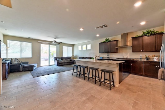 kitchen featuring a center island with sink, stovetop, a kitchen breakfast bar, wall chimney range hood, and dark brown cabinetry