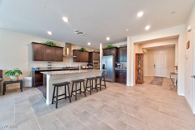 kitchen with an island with sink, stainless steel appliances, a breakfast bar, dark brown cabinets, and wall chimney range hood