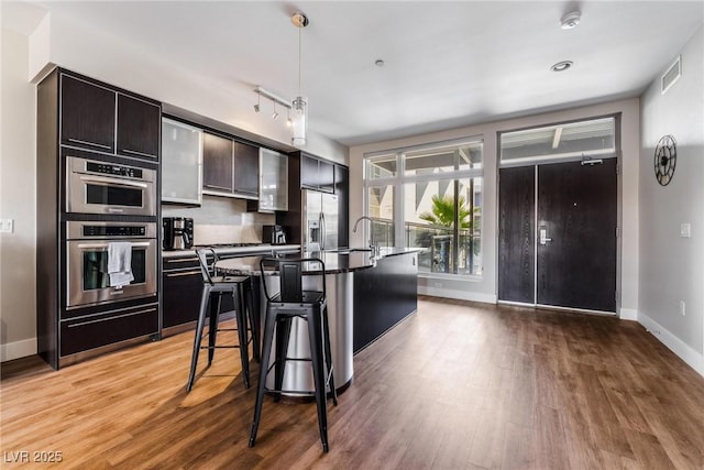 kitchen with a kitchen island with sink, stainless steel appliances, dark brown cabinetry, light hardwood / wood-style flooring, and a breakfast bar area