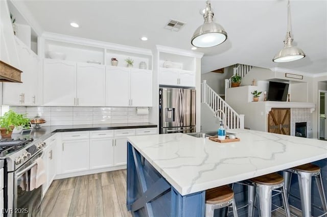 kitchen featuring a center island with sink, hanging light fixtures, stainless steel appliances, dark stone counters, and white cabinets