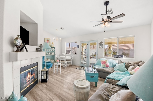 living room with french doors, a tiled fireplace, lofted ceiling, light hardwood / wood-style flooring, and ceiling fan with notable chandelier