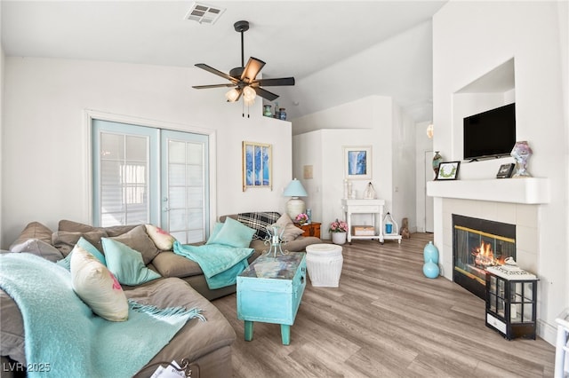 living room featuring lofted ceiling, a tile fireplace, light hardwood / wood-style floors, ceiling fan, and french doors