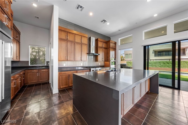 kitchen featuring wall chimney range hood, range, a kitchen island with sink, and tasteful backsplash