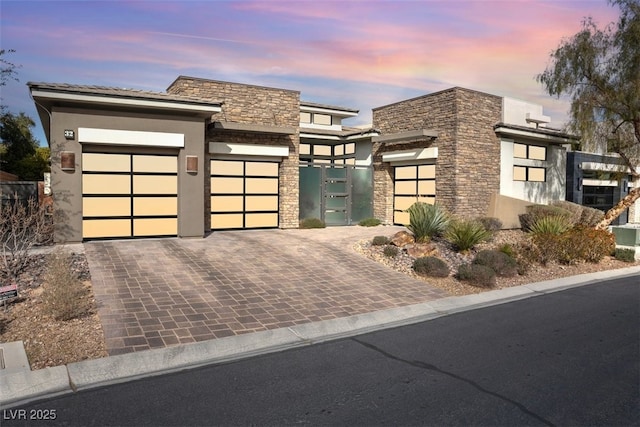 modern home with a garage, stone siding, decorative driveway, and stucco siding