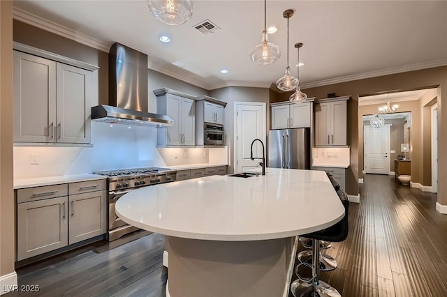 kitchen with appliances with stainless steel finishes, sink, decorative light fixtures, wall chimney range hood, and an island with sink