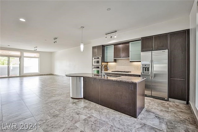 kitchen featuring a kitchen island with sink, stainless steel appliances, dark brown cabinetry, light stone countertops, and sink