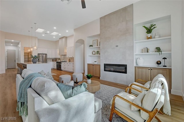 living room with ceiling fan, light wood-type flooring, a tile fireplace, and built in features