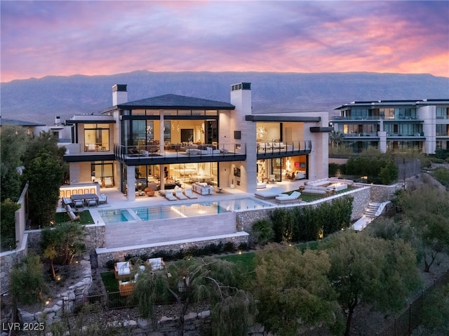 back house at dusk with a mountain view, a balcony, a patio area, an in ground hot tub, and an outdoor living space