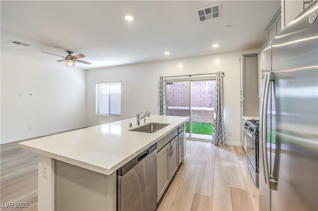 kitchen with light wood-type flooring, a center island with sink, gray cabinetry, sink, and appliances with stainless steel finishes