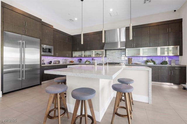 kitchen with built in appliances, a breakfast bar area, wall chimney exhaust hood, tasteful backsplash, and decorative light fixtures