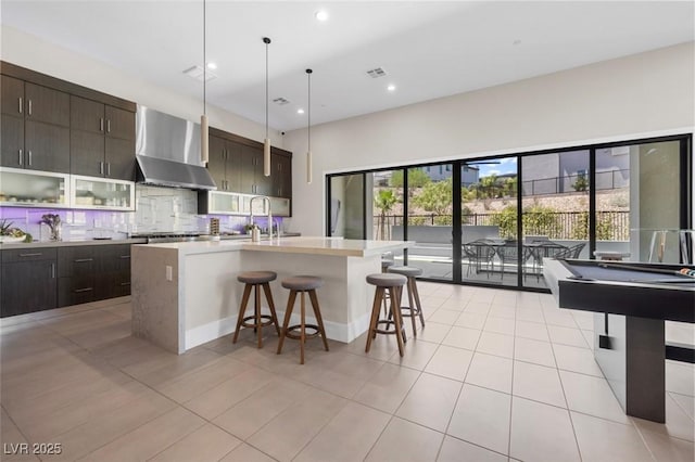 kitchen featuring light tile patterned floors, a breakfast bar, decorative backsplash, wall chimney exhaust hood, and a center island with sink