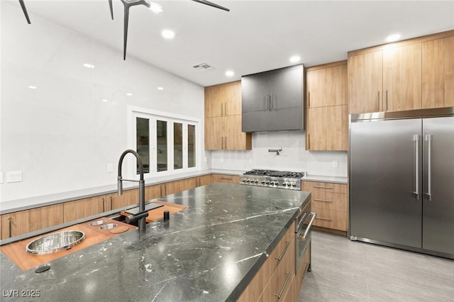 kitchen with dark stone counters, stove, stainless steel built in refrigerator, and wall chimney exhaust hood