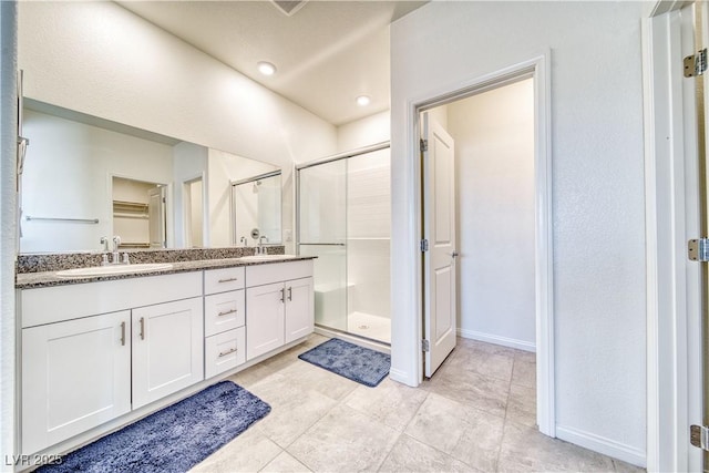 full bathroom with a walk in closet, double vanity, a sink, a shower stall, and baseboards