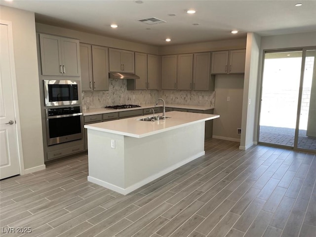 kitchen featuring appliances with stainless steel finishes, gray cabinetry, an island with sink, decorative backsplash, and sink