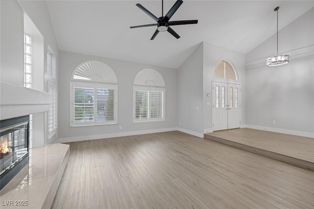 unfurnished living room featuring a premium fireplace, wood finished floors, high vaulted ceiling, baseboards, and ceiling fan with notable chandelier