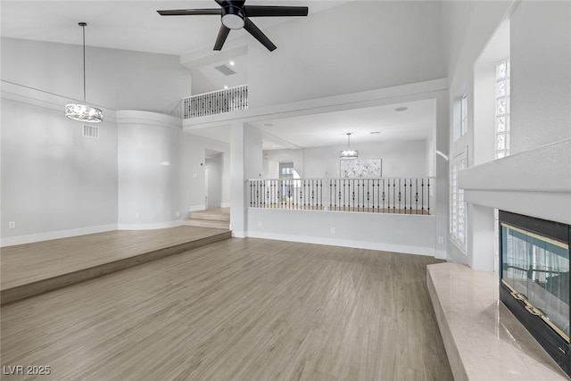 unfurnished living room with baseboards, a glass covered fireplace, a towering ceiling, wood finished floors, and ceiling fan with notable chandelier